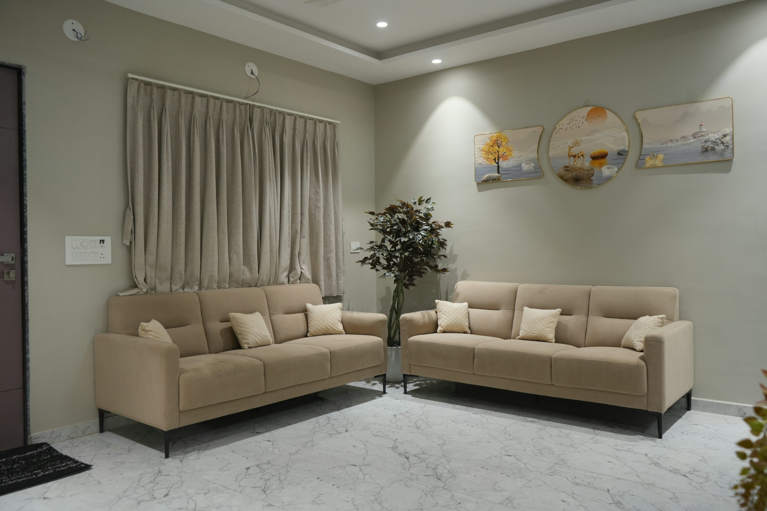 Due divani beige con cuscini sono disposti ad angolo in un soggiorno con tende chiare e quadri alle pareti.