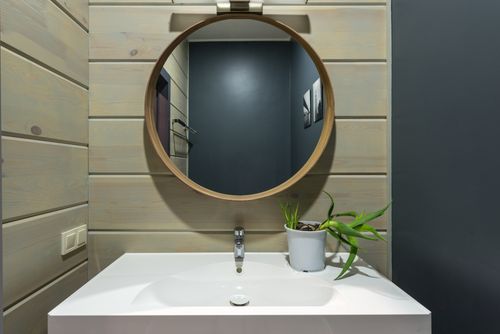 Specchiera rotonda sopra lavabo bianco; pareti in legno chiaro e muro blu scuro sullo sfondo; pianta in vaso grigio accanto al rubinetto.