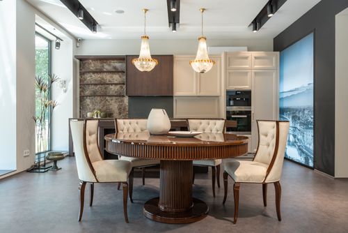 Immagine di sala da pranzo con arredamento classico: tavolo rotondo in legno, sei sedie imbottite crema, lampadari d’epoca e cucina moderna sullo sfondo, tono elegante.