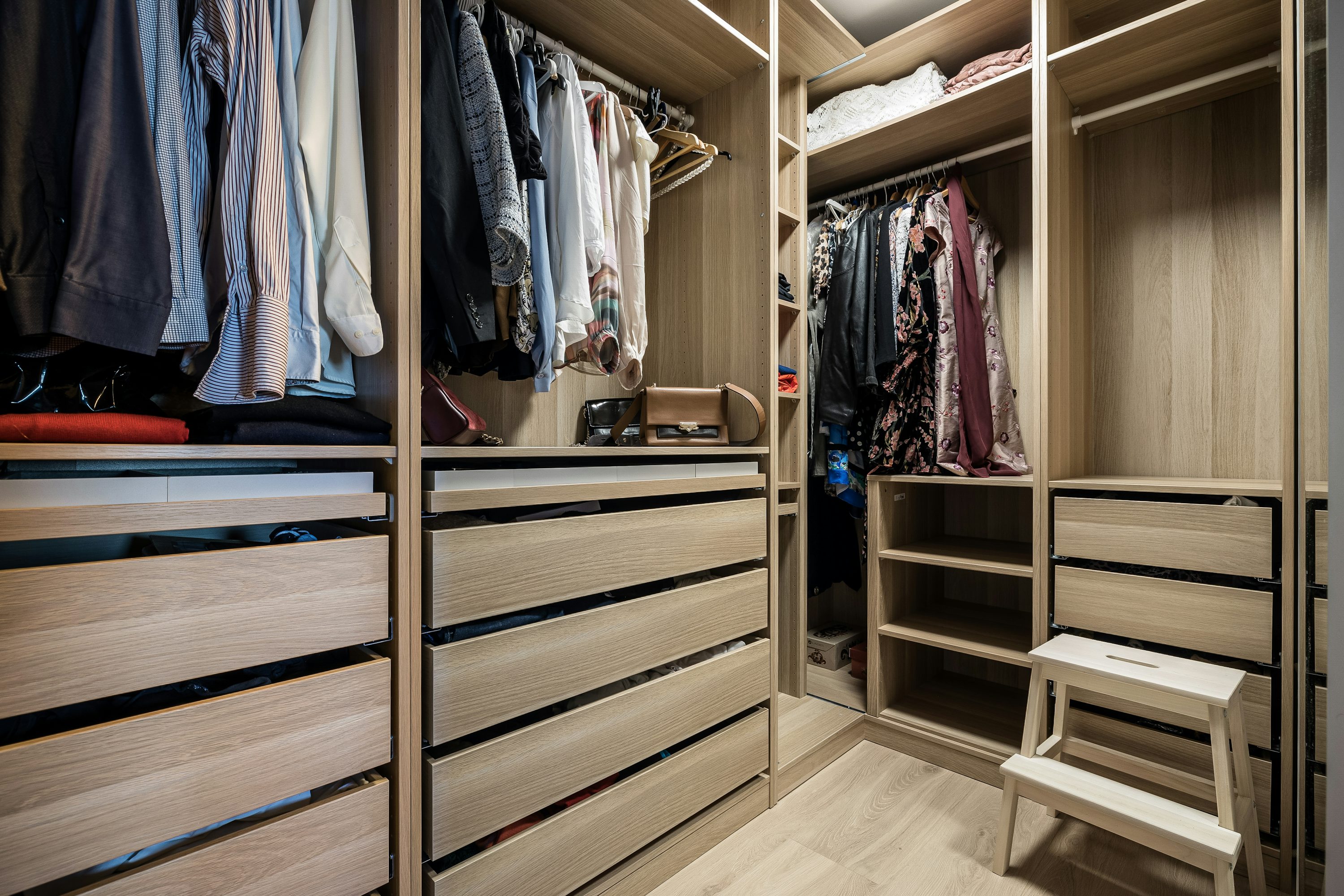 Interna di cabina armadio in stile moderno con ante tirature e cassetti in legno chiaro, abiti appesi, accessori e scaffali.