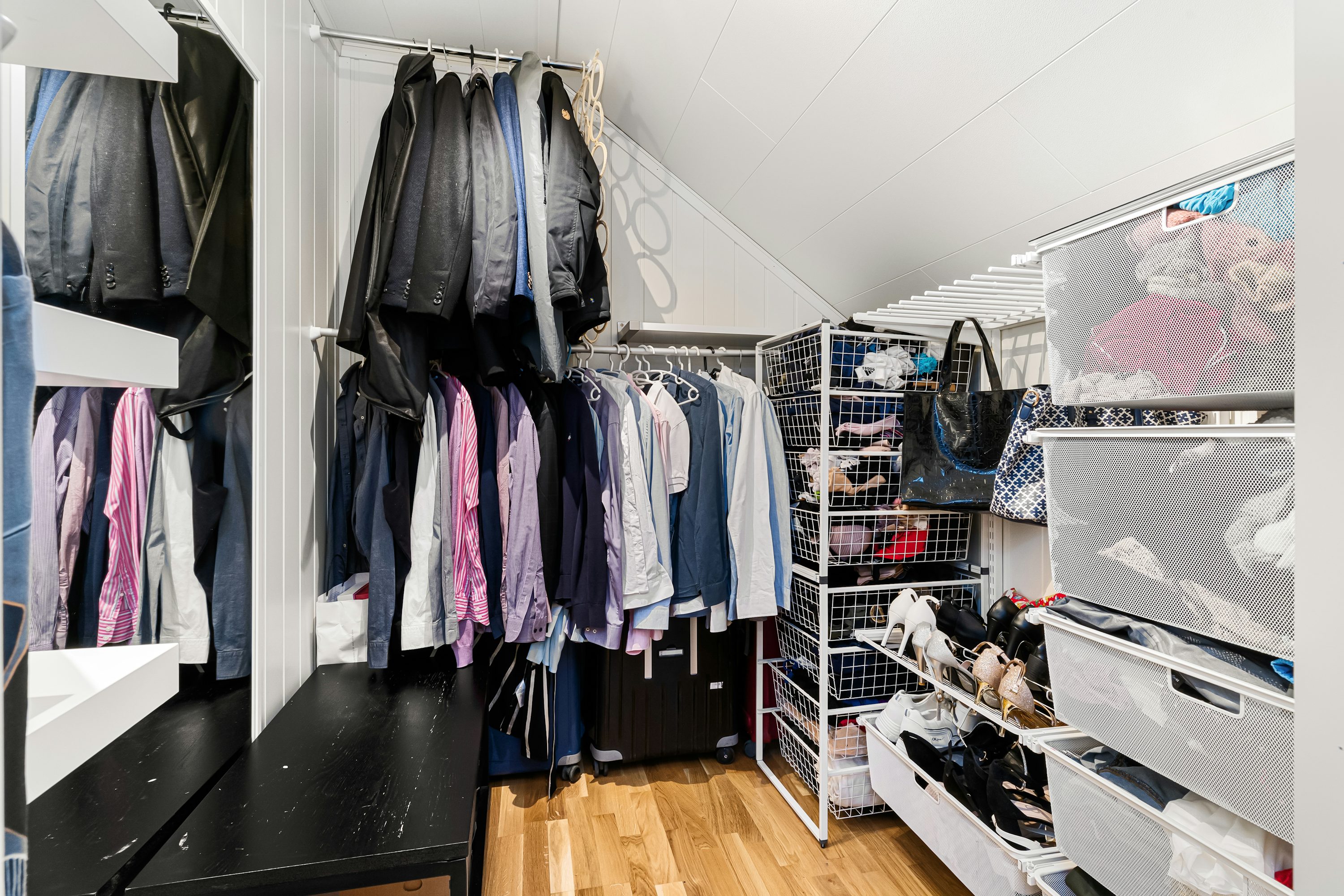 Cappotti e camicie appese lungo parete e scatole contenenti scarpe e accessori in cabina armadio luminosa e ben organizzata.